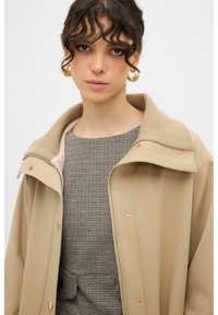Beige jas met een hoge kraag en drukknoopsluiting. Draag over een gemusterde, geruite top. Soepele textuur met contrasterende stofdetails.
