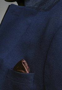 Granatowa marynarka z teksturowanej tkaniny; posiada poszetkę w brązowym jedwabiu z subtelnym wzorem. Widać czyste linie i dopasowany krój.