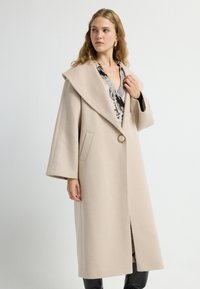 Beige uldblandede frakke med bred krave, lange ærmer, frontlukning med knapper, sidelommer og lige snit, der har kontrasterende søm.