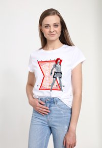 Hvid bomulds T-shirt med et grafisk print af en karakter med rødt hår og en grå dragt, sat mod en rød og tekstureret baggrund.