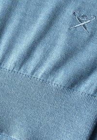Tissu tricoté bleu clair avec ourlet côtelé et petit détail brodé de deux aiguilles à tricoter croisées près du coin supérieur droit.