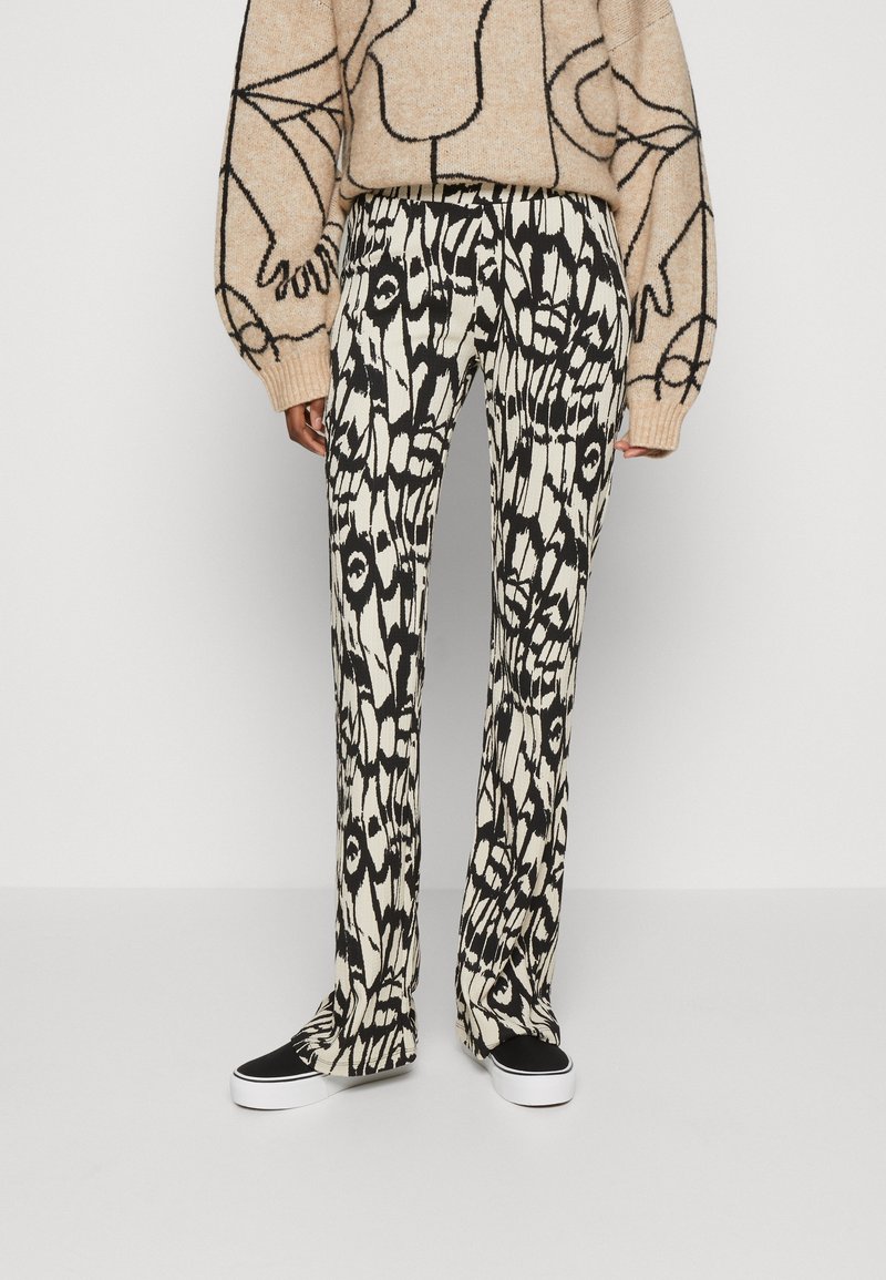 Pantalon large avec un motif abstrait noir et blanc, porté avec des chaussures slip-on noires et blanches et un pull beige avec un dessin au trait noir.