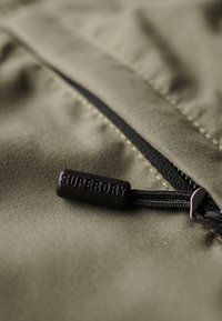 Tessuto verde oliva con una cerniera nera dotata di un pull tab testurizzato contrassegnato "SUPERDRY". Cuciture dettagliate visibili lungo la linea della cerniera.