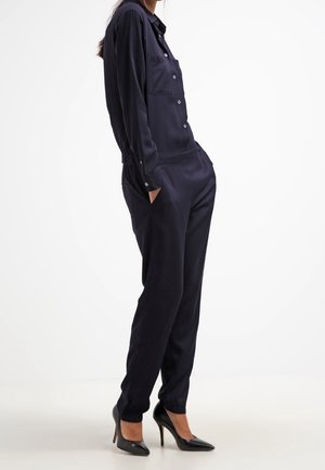Marineblauw jumpsuit gemaakt van gladde stof, met een knoopsluiting aan de voorkant, lange mouwen, twee borstzakken en taps toelopende pijpen. Draagt zwarte hakken.