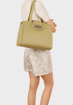Donna che indossa una gonna floreale color crema e un maglione chiaro, con una grande borsa di pelle verde oliva sulla spalla.