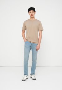 Νεαρός άνδρας που στέκεται με το ένα χέρι στην τσέπη ενός ανοιχτομπλε τζιν, φορώντας μπεζ μπλουζάκι και λευκά αθλητικά παπούτσια με μαύρες ρίγες μπροστά σε λευκό φόντο.