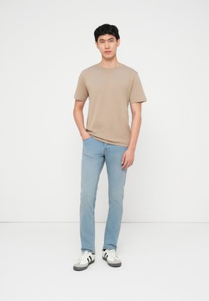 Tânăr în picioare cu o mână în buzunarul blugilor deschis la culoare, purtând un tricou bej și adidași albi cu dungi negre, pe un fundal alb.