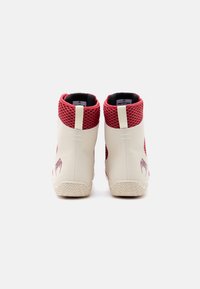 Ζεύγος μπότες υψηλού κορμού με μπεζ σόλες και σώματα, κόκκινες δίχτυες περιμετρικές στηρίξεις αστραγάλου και ένα μικρό σκούρο κόκκινο γραφικό στη φτέρνα, εμφανιζόμενα από πίσω.