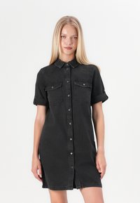 Zwarte denim shirt jurk met korte mouwen, knopen aan de voorkant, twee borstzakken en een lengte tot half boven de dij. Voorzien van een kraag en opgerolde manchetten.