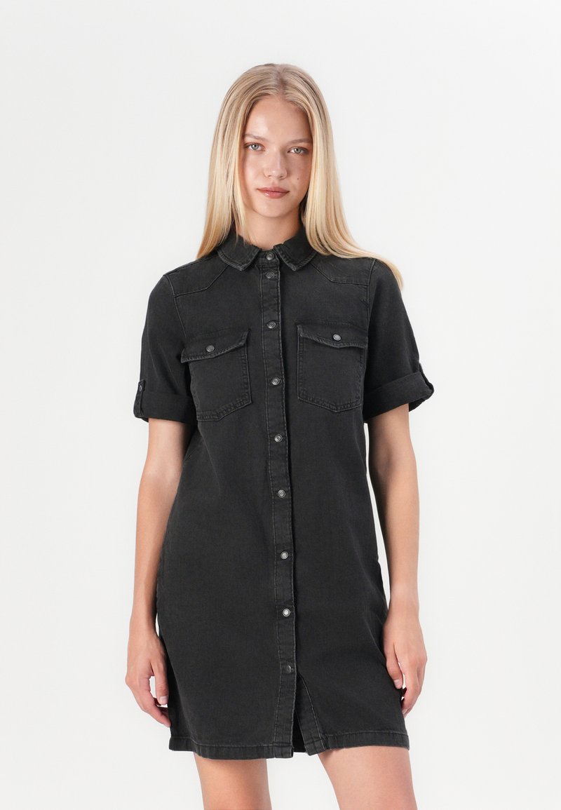 Zwarte denim shirt jurk met korte mouwen, knopen aan de voorkant, twee borstzakken en een lengte tot half boven de dij. Voorzien van een kraag en opgerolde manchetten.
