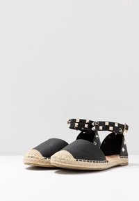 Espadrilles de cuero negro con punteras trenzadas en beige y tiras al tobillo adornadas con tachuelas doradas en una superficie blanca.