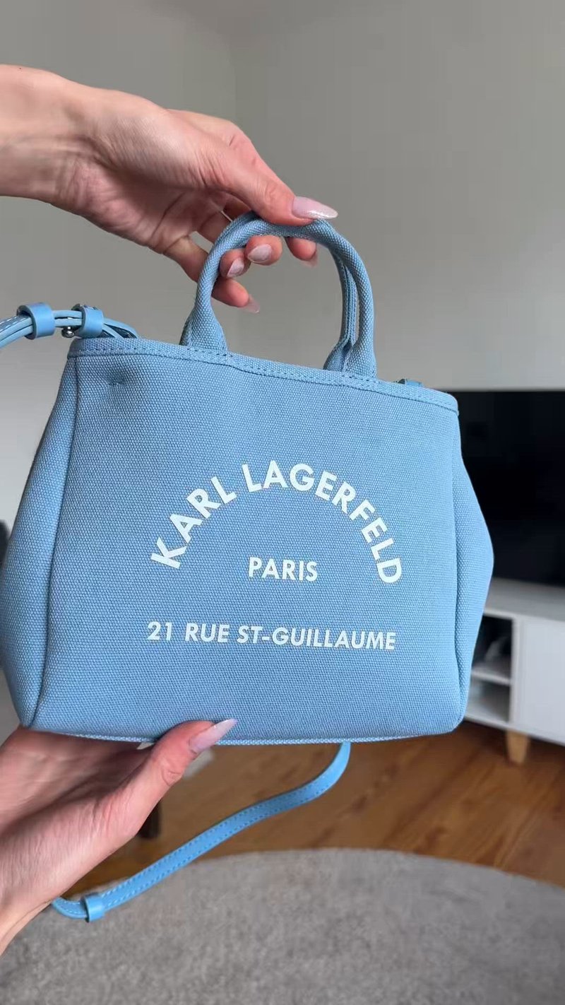 Ръка държи малка синя чанта Karl Lagerfeld Paris с горни дръжки и бял текст, на закрито с телевизор и шкаф на заден план.