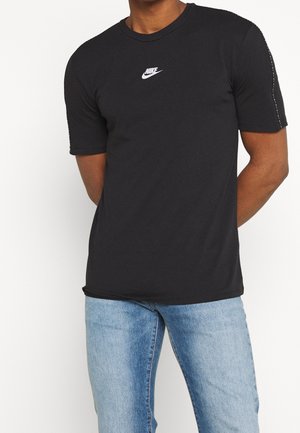 Mężczyzna w czarnej koszulce Nike z małym białym logo na piersi i jasnoniebieskich jeansach, stoi z rękami za plecami.
