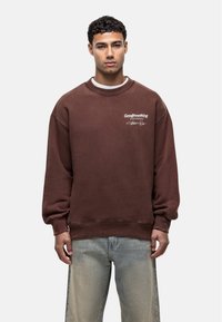 Felpa marrone con scollo rotondo e polsini a costine. Presenta il logo bianco "Goodfornothing" e dettagli caratteristici. Indossata con jeans azzurri chiari.