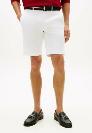 Mann trägt weiße Shorts, schwarze Loafers, einen schwarzen Gürtel mit roten und weißen Streifen und ein rotes Hemd, steht mit einer Hand in der Tasche.
