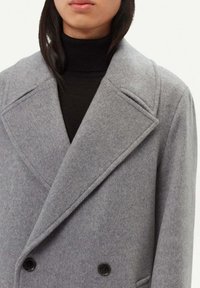 Grå dubbelknäppt kappa i ull med spetsig slag, utrustad med svarta knappar och en mjuk, texturerad yta. Bärs över en svart polotröja.