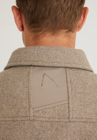 Brun herringbone-tygkrage med en beige lapp och broderade detaljer. Kragen har en strukturerad design och ren sömnad.