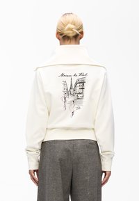 White cropped sweatshirt with a folded collar, featuring a black and pink cityscape illustration and "Maison de Kol" embroidered on the back.