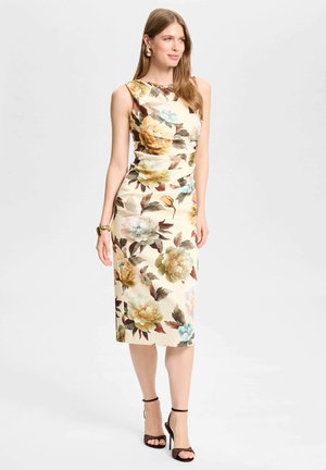 Vrouw staand, met een mouwloze beige midi-jurk met grote bloemenprint en zwarte hoge hakken met enkelbandje tegen een effen achtergrond.