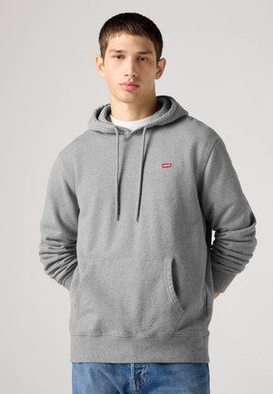 Νέος άνδρας φοράει γκρι φούτερ Levi's με τσέπη μπροστά και κορδόνια, στέκεται μπροστά σε απλό φωτεινό φόντο.