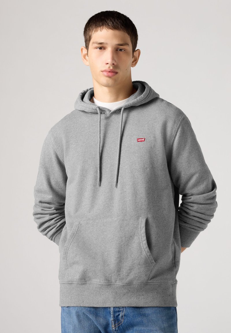 Νέος άνδρας φοράει γκρι φούτερ Levi's με τσέπη μπροστά και κορδόνια, στέκεται μπροστά σε απλό φωτεινό φόντο.