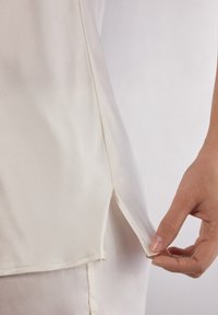 Silkblus i off-white med en mjuk textur; har en slits vid fållen och subtila sömdetaljer; hålls av en hand.