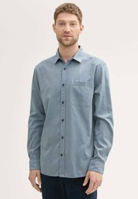 Camisa abotonada de color azul claro con un sutil patrón geométrico, de mangas largas, un bolsillo en el pecho y botones negros. Textura de tela suave.