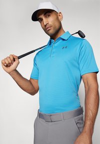 Camisa de polo azul con tejido texturizado, mangas cortas y un cuello con tres botones, combinada con pantalones grises y una gorra blanca. Sosteniendo un palo de golf de manera casual.
