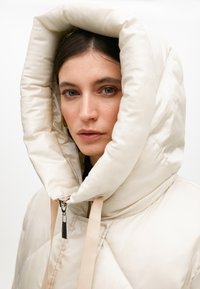 Puffig krämfärgad jacka med en stor, strukturerad huva. Har en dragkedjestängning och beige remmar. Slät, glänsande textur med diamant-quiltmönster.