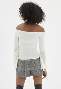 Weißes Top mit schulterfreiem Design und strukturierten, glockenförmigen Ärmeln sowie taillierter Passform. Kombiniert mit grauen Karo-Shorts, die einen lässigen Stil zeigen.