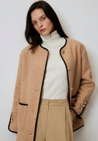 Cappotto in misto lana beige con rifiniture nere, dotato di colletto rotondo, tasche frontali e dettagli in bottone dorato. Indossato sopra un dolcevita bianco.