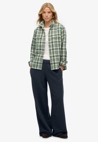 Groene en witte geruite flanellen shirt met knoopsluiting, gedragen over een wit shirt. Gecombineerd met donkerblauwe wijde broeken en zwarte schoenen.