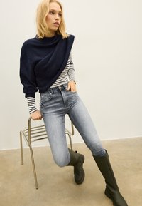 Mörkblå pullover i ullblandning över en randig långärmad topp, kombinerad med höga ljusblå jeans och mörkgröna gummistövlar.