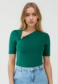Model met een groene, aansluitende top met een draai-detail en een uitsnijding, gecombineerd met hoge denim shorts, tegen een effen achtergrond.