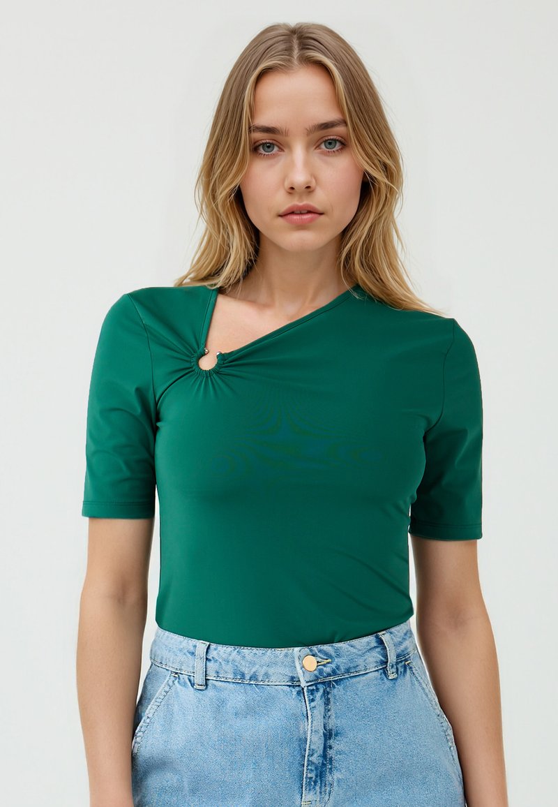 Model met een groene, aansluitende top met een draai-detail en een uitsnijding, gecombineerd met hoge denim shorts, tegen een effen achtergrond.