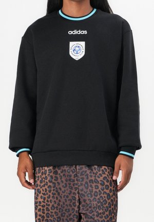 Personne portant un sweat Adidas noir avec un col et des poignets bordés de bleu, et un pantalon marron à imprimé léopard.