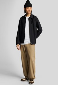 Chaqueta negra de botones sobre una camiseta blanca, combinada con pantalones beige de pierna ancha. Un gorro oscuro y zapatillas marrones con estampado completan el look.