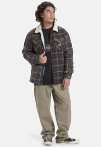 Chaqueta de franela a cuadros en marrón y gris con un cuello crema, cierre de cremallera, dos bolsillos en el pecho y un parche de la marca. Combinada con pantalones sueltos de color beige.