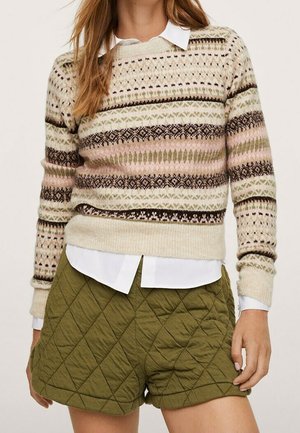 Kobieta ubrana w kremowy, różowy i brązowy wzorzysty sweter na białej koszuli z kołnierzykiem, w połączeniu z oliwkowozielonymi pikowanymi szortami.