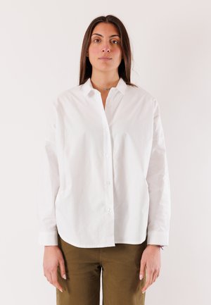 Giovane donna con lunghi capelli scuri indossa una camicia bianca abbottonata e pantaloni verde oliva, in piedi davanti a uno sfondo chiaro e semplice.