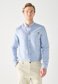 Light blue button-up shirt with long sleeves, chest pocket, and brown buttons. Soft fabric texture, tailored fit, complemented by beige trousers.