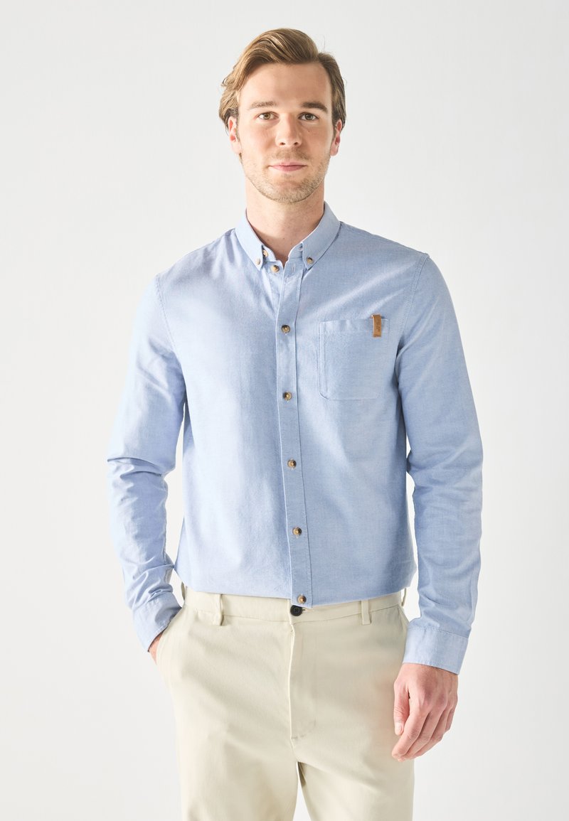 Light blue button-up shirt with long sleeves, chest pocket, and brown buttons. Soft fabric texture, tailored fit, complemented by beige trousers.