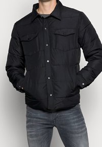 Man som bär en svart knappad vadderad jacka med fickor på bröstet och händerna i sidofickorna, i kombination med blekta grå jeans.