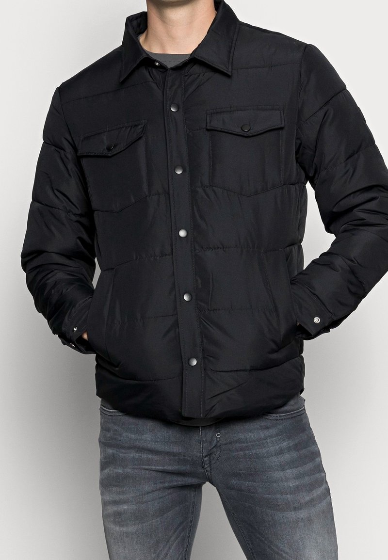 Man som bär en svart knappad vadderad jacka med fickor på bröstet och händerna i sidofickorna, i kombination med blekta grå jeans.