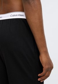Μαύρο βαμβακερό σορτς με λευκή ελαστική μέση που φέρει το κείμενο «Calvin Klein». Λεία υφή, εφαρμοστή γραμμή, εμφανίζεται από το πλάι.