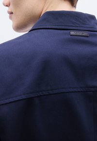 Μπλε μπουφάν ναυτικού με λεία υφή, που διαθέτει κλασικό γιακά και μικρή ετικέτα με το λογότυπο στο πίσω μέρος. Απλός, δομημένος σχεδιασμός.