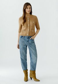 Beiger Strickcardigan mit kurzen Ärmeln, Fronttaschen und dekorativen Knöpfen, kombiniert mit hellblauen lockeren Jeans und gelben Knöchelschuhen.