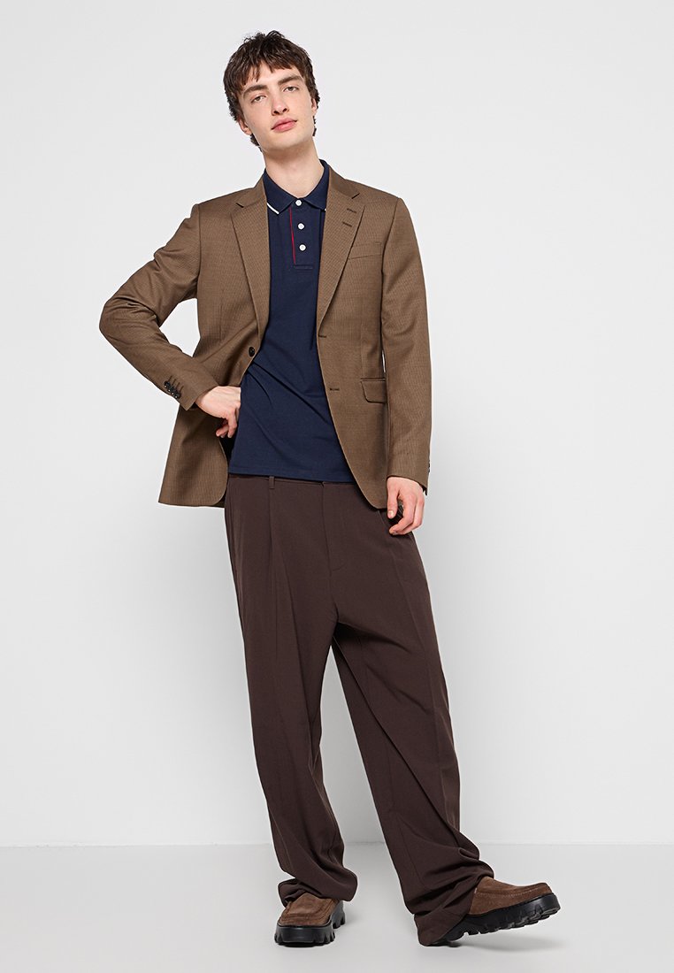Blazer de cuadros marrón, camiseta polo azul marino y pantalones de pierna ancha marrones. Textura suave, corte ajustado y calzado oscuro con suela gruesa.