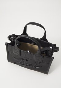 Černá kožená kabelka s texturovaným povrchem, vybavená krátkými rukojeťmi a viditelným logem na přední straně. Interiér je v světlejší neutrální barvě.