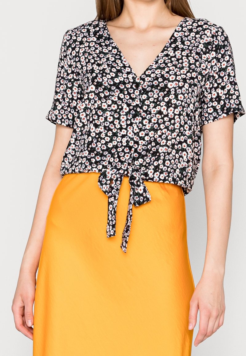 Frau in einer schwarzen kurzärmligen Bluse mit weißem und rotem Blumenmuster, die an der Taille gebunden ist, und einem hochgeschnittenen leuchtend orangefarbenen Rock.